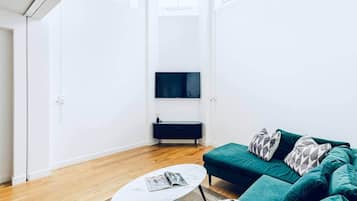 A modern living room with a flat-screen TV, a dark console table, a teal sofa with patterned cushions, and a small round table with a magazine.