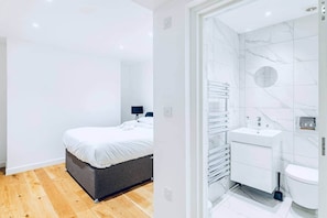 A modern bedroom with a bed, bedside table, and lamp, and a bathroom with a sink, toilet, and towel rack. - Detached Family Home with Free Parking (London)