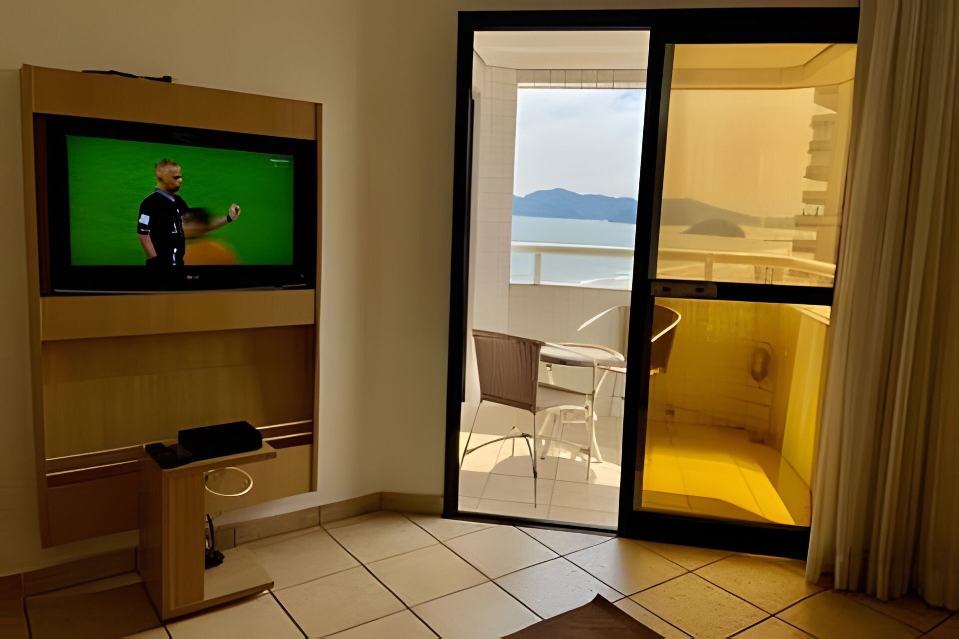 Una habitación moderna con una televisión en la pared, una pequeña mesa con sillas, y vistas a un balcón con piscina y montañas al fondo.