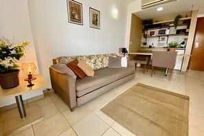 A modern living room with a sofa, a dining table, and a kitchen area visible through an open doorway. - SHB Charmoso Flat Com Vista Mar (Santos)