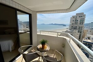 A balcony with a view of the beach and a tall building. There is a bed and a small table with a plant. - SHB Charmoso Flat Com Vista Mar (Santos)