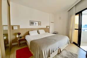 Quarto de hotel com cama, mesa de cabeceira, tapete vermelho, quadro na parede e vista para o exterior.