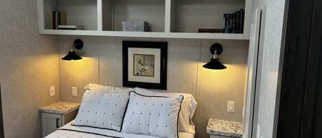 A bedroom with a bed, two bedside tables, a nightstand, a shelf above the bed, and two wall-mounted lamps.