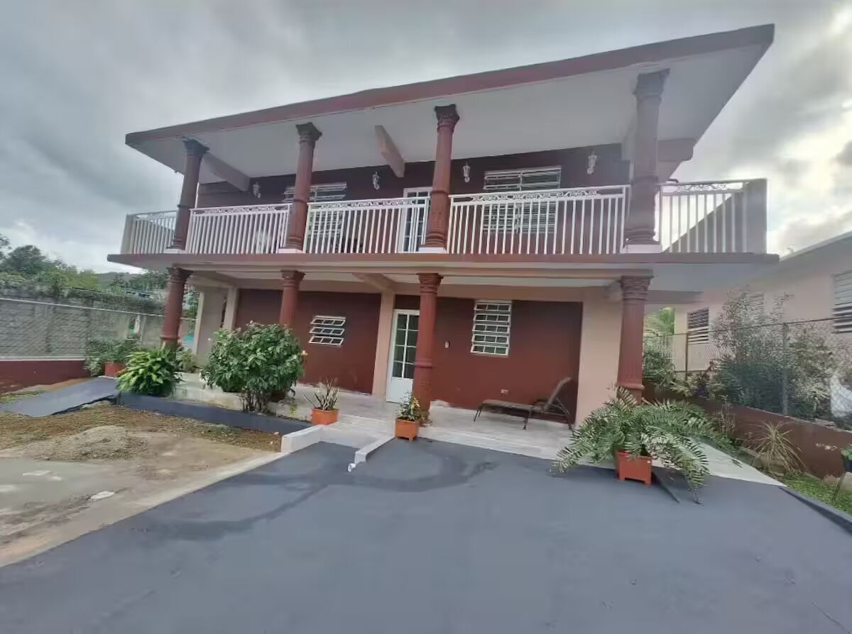 A two-story building with a balcony, potted plants, and a paved entrance area.