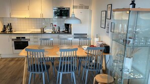 Ett modernt kök med vita skåp, en inbyggd ugn, en mikrovågsugn och en kaffebryggare. Här finns ett matbord med stolar och ett glasskåp med vinglas.