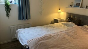 Ett sovrum med en säng, ett nattduksbord, en lampa, ett element, ett fönster med gardiner, en växt och en tavla på väggen.