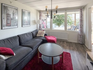 Una sala moderna con un sofá gris, una mesa redonda y un tapete rojo. Hay dos cuadros en la pared, una ventana con cortinas y un radiador.