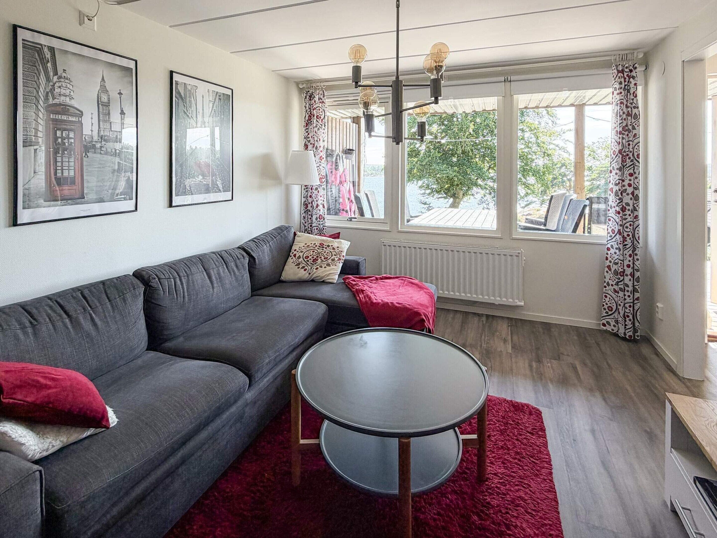 A modern living room with a grey sofa, a round table, and a red rug. There are two framed pictures on the wall, a window with curtains, and a radiator.