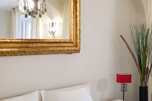 Ein Zimmer mit einem großen, verzierten Spiegel, einem weißen Bett, einer roten Lampe und einer hohen Pflanze.