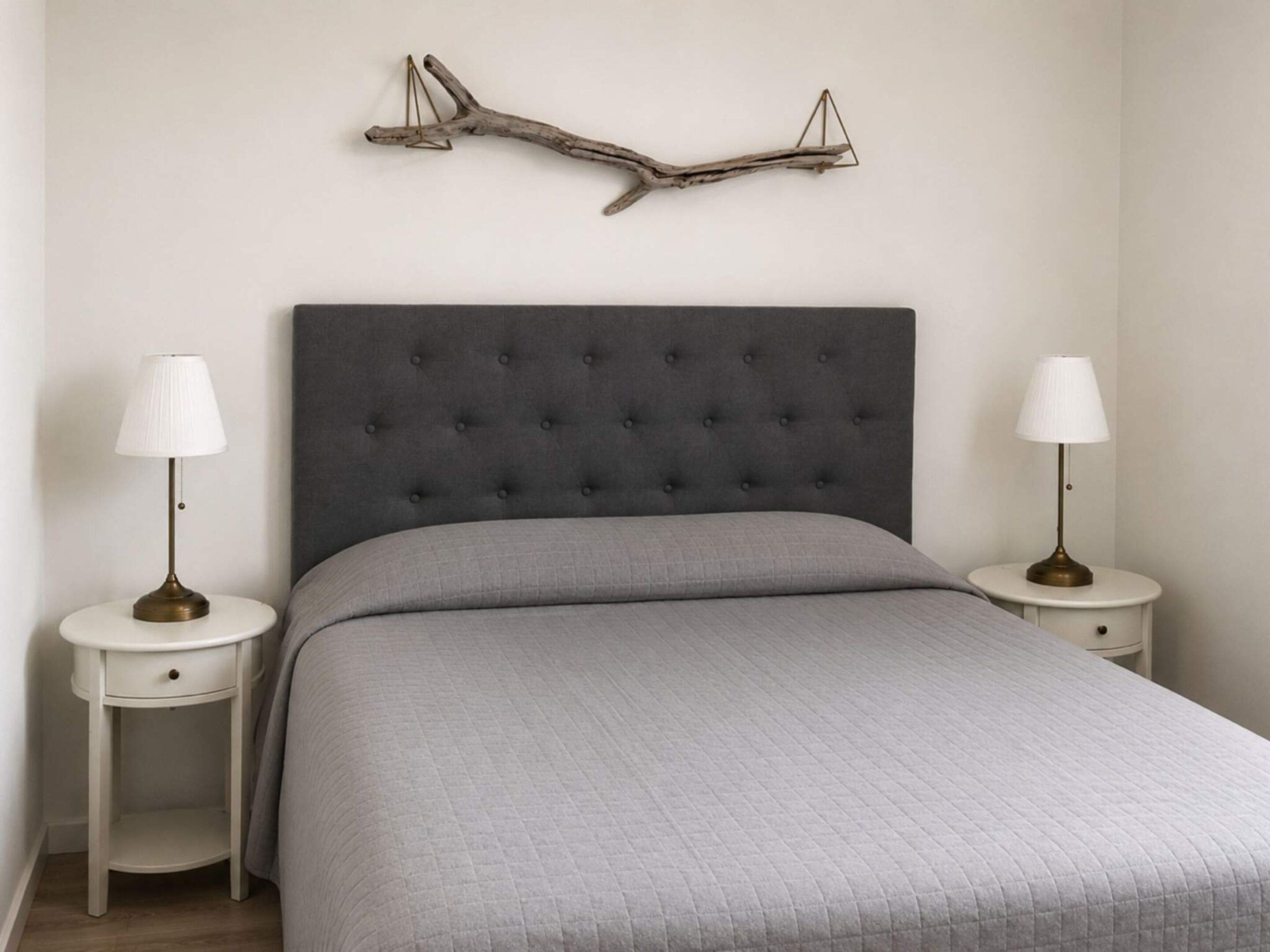 Quarto com cabeceira estofada, colcha acolchoada cinza e duas mesas de cabeceira com abajures.