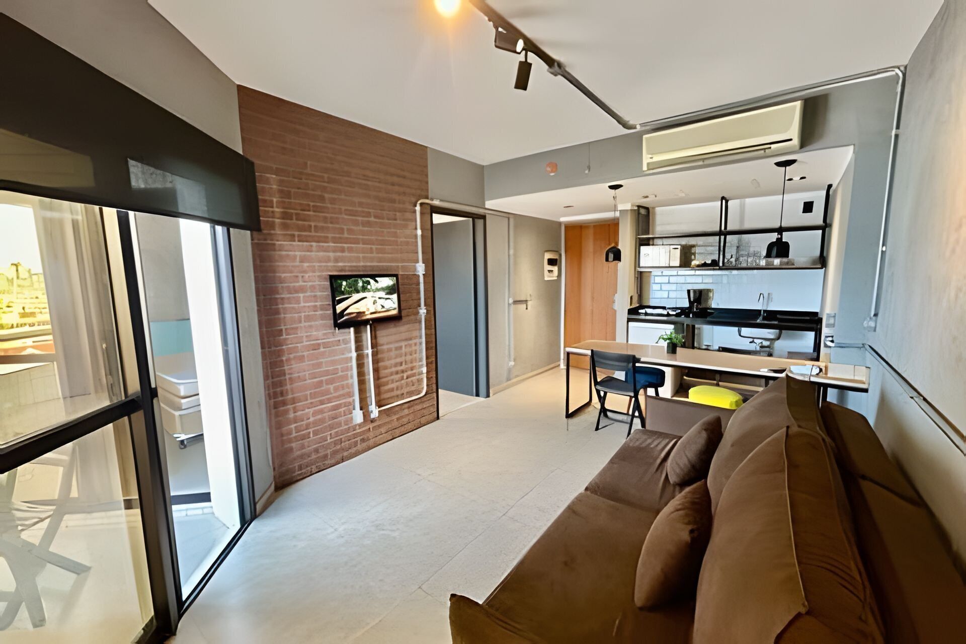 Una sala de estar moderna con un sofá marrón, un televisor montado en la pared de ladrillo y una zona de cocina con armarios y encimera.