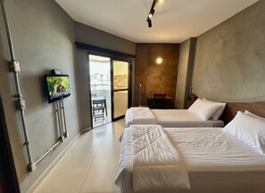 A hotel room with two beds, a TV mounted on the wall, a chair, and a view of the city through a glass door. - SHB Desfrute do Conforto à Beira-Mar (Santos)