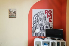 A room with a TV on a white stand, a poster of the Colosseum, and a small table with a lamp and decorative items. - Villa Borghese House (Rome)