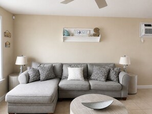 A neutral-toned living room with a grey sectional sofa, patterned throw pillows, two side tables with lamps, a decorative shelf with a 'HOME' sign, and a round coffee table.