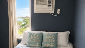 A bed with a striped blue and white comforter, two pillows with text, a window with curtains, and a wall-mounted air conditioning unit. - Waterfront Lagoon Escape , Sunset Views, Private Dock and Minutes to LBI (Stafford Township)