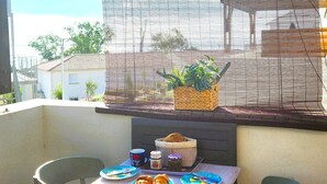 Un balcón con una mesa lista para dos personas, con platos de comida, una cesta con una planta y una pequeña mesa con una taza y un tarro.