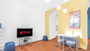 Un salon moderne doté d’un téléviseur à écran plat, d’une table à manger avec des chaises, d’un pouf bleu et d’un tableau abstrait aux couleurs vives accroché au mur.
