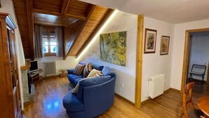 A cozy living room with a blue sofa, wooden beams, a painting, and a dining area. - Benasque Shelter (Benasque)