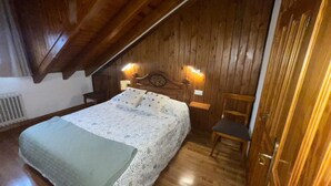 A cozy bedroom with a wooden ceiling, a bed with a floral bedspread, two wall-mounted lights, and a wooden chair. - Benasque Shelter (Benasque)