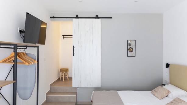 Un dormitorio con cama, televisor montado en la pared, perchero de madera y una escalera que lleva a otra habitación.