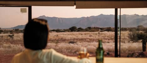 Una persona sentada a una mesa con una botella y un vaso, con vista a un paisaje desértico y montañas al fondo.