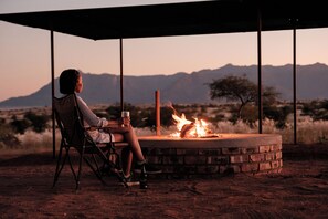 A person seated at a table with a fire pit, under a shelter, with a mountainous landscape in the background.