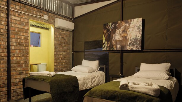 A hotel room with two beds, a brick wall, an air conditioning unit, and a painting on the wall.