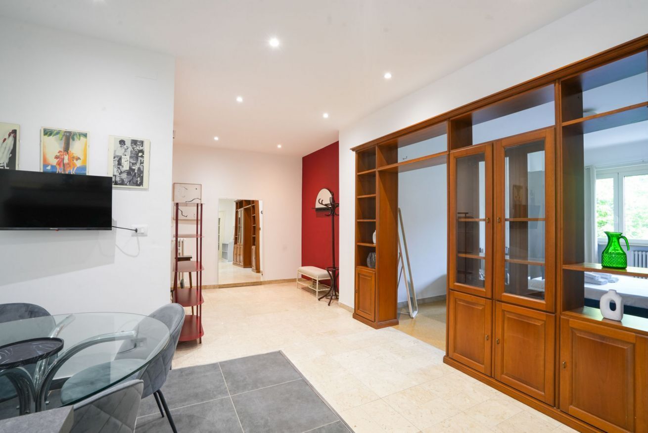 A modern living room with a glass table, a red accent wall, and a wooden cabinet with glass doors.