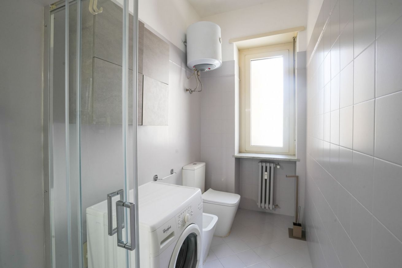 A compact bathroom with a glass shower enclosure, a washing machine, a toilet, and a radiator.