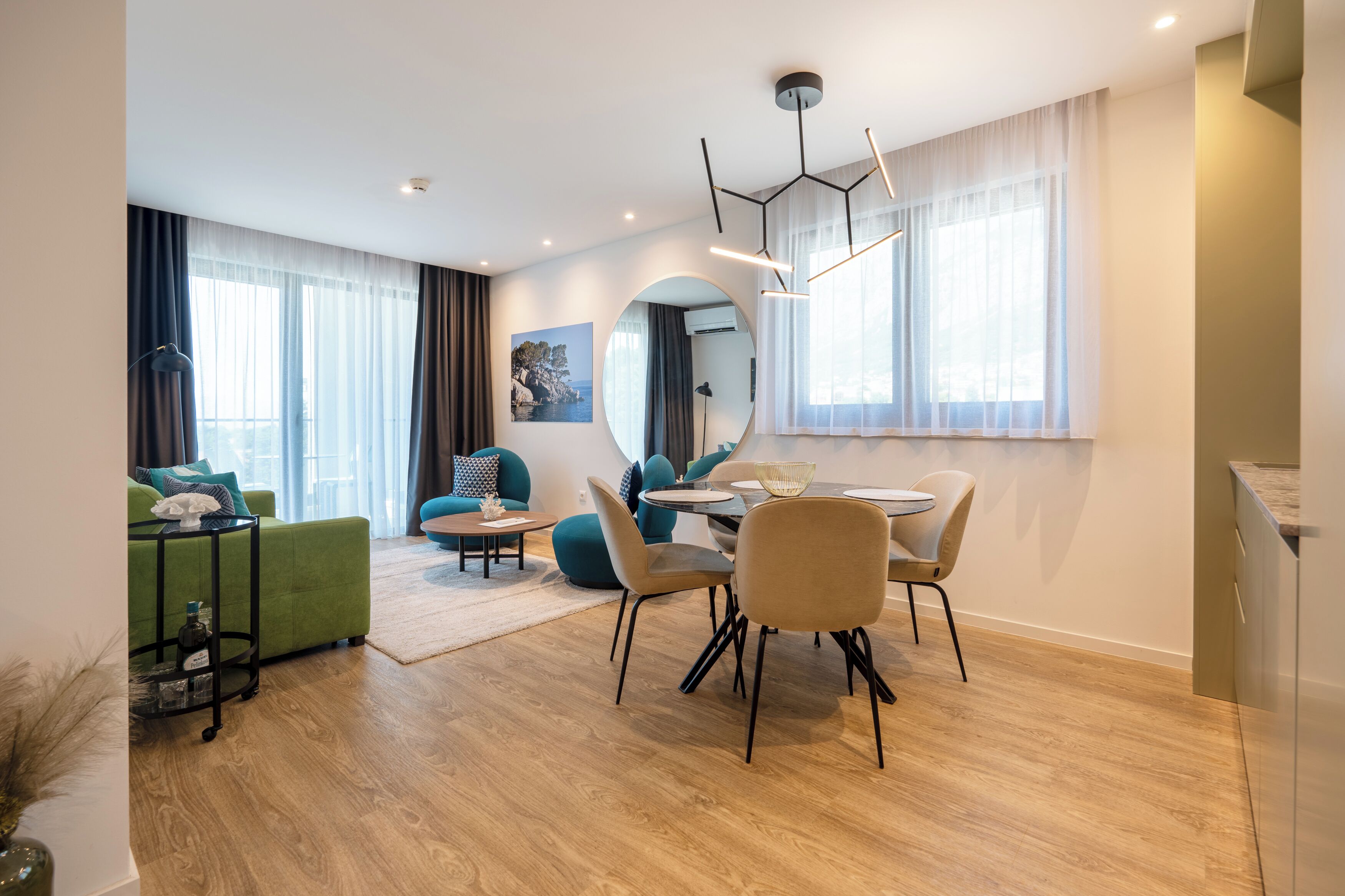 Ein modernes Wohnzimmer mit einem grünen Sofa, einem Esstisch mit Stühlen, einem runden Spiegel und einem großen Fenster mit Vorhängen.