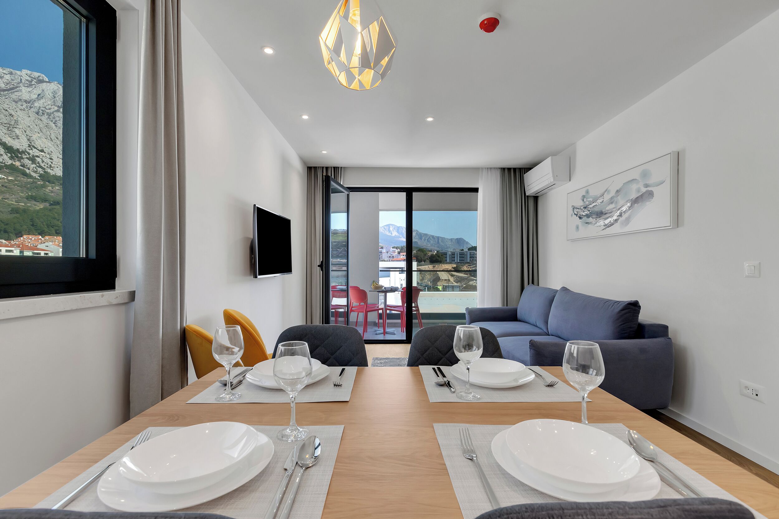 A modern hotel room with a dining table set for two, a sofa, a TV, and a balcony with a mountain view.