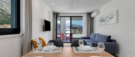 A modern hotel room with a dining table set for two, a sofa, a TV, and a balcony with a mountain view.