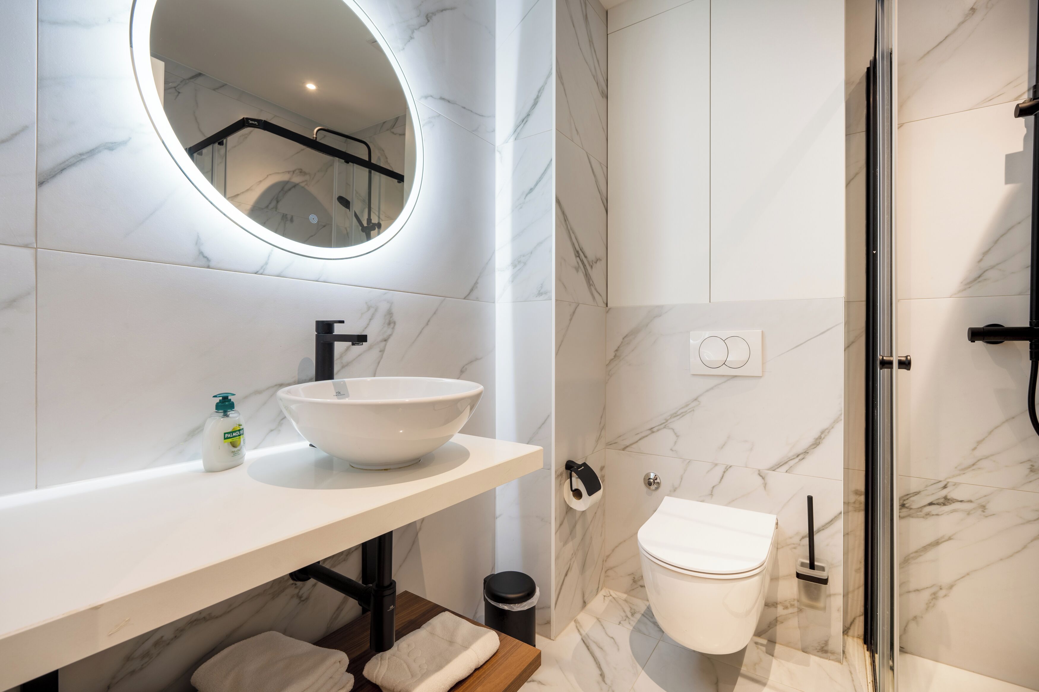 A modern bathroom with a white marble sink, a round mirror, a toilet, and a glass shower enclosure.