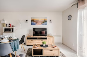 A modern living room with a flat-screen TV, wooden furniture, a dining area with a table set for two, and a painting on the wall. - Cozy Studio - CDG - Exhibition Center - ARENA Grand Paris (Sevran)