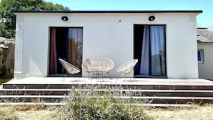 Een modern gebouw met twee openslaande deuren, een klein terras met twee stoelen, en een heldere lucht.