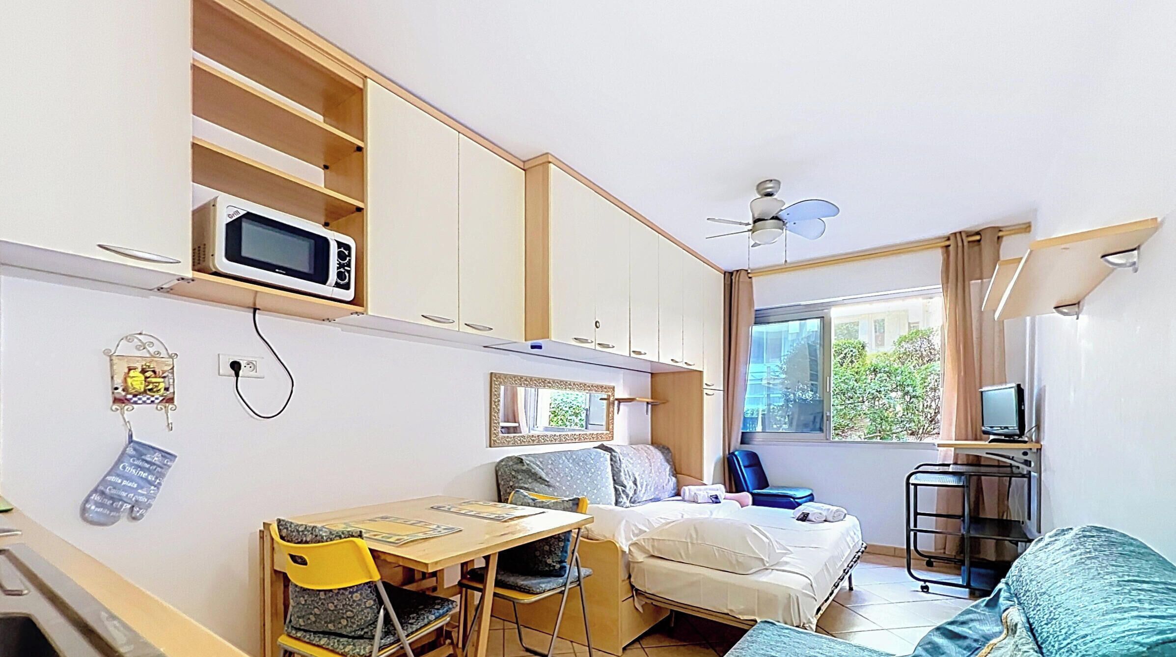 A compact room with a bed, a desk with a chair, a microwave, a ceiling fan, and a window with a view of greenery.