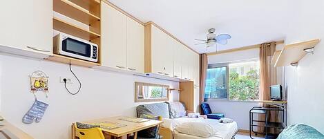 A compact room with a bed, a desk with a chair, a microwave, a ceiling fan, and a window with a view of greenery.