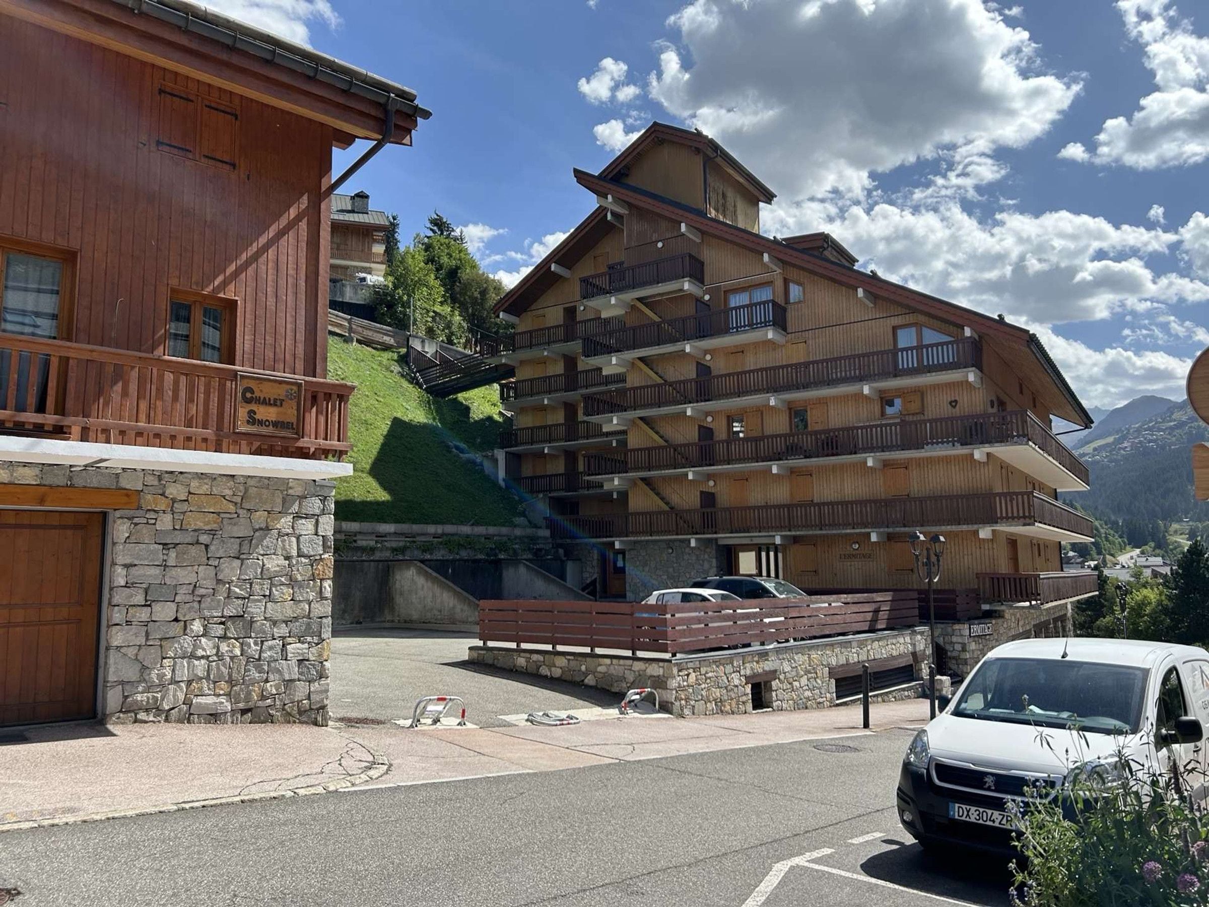 A multi-story wooden building with balconies, a stone facade, and a parking area with a white van.