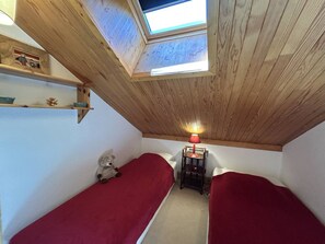 Ein kleines Zimmer mit zwei Einzelbetten, einem Oberlicht, einem Tisch mit Lampe und einem Teddybär.