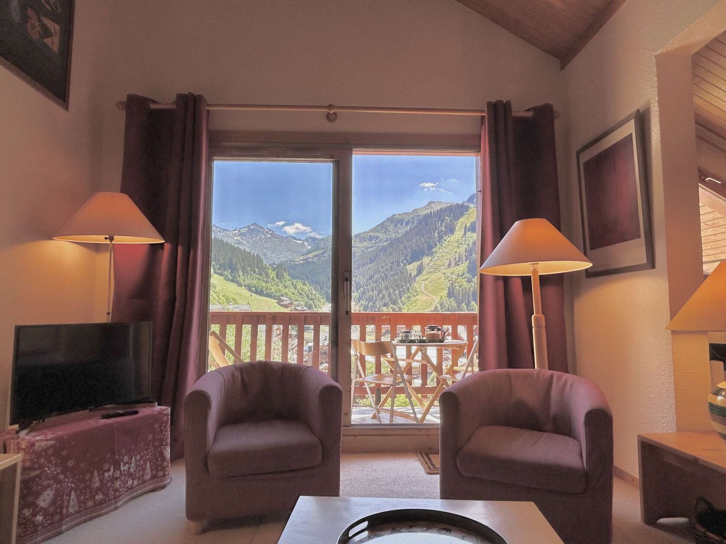 A room with a TV, two armchairs, a table, and a balcony with a mountain view.