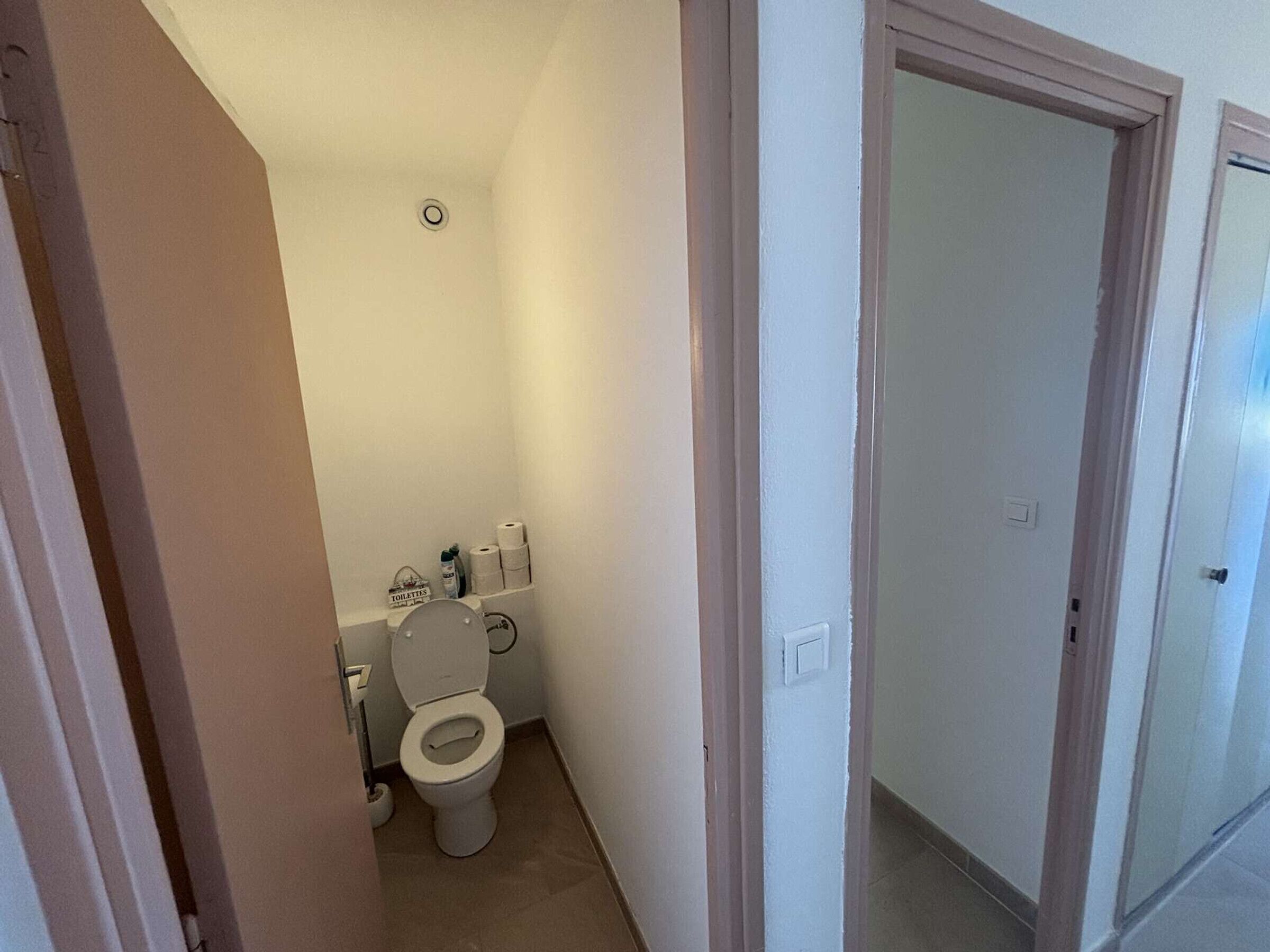A small, narrow bathroom with a toilet, a roll of toilet paper, and a small shelf with toiletries.