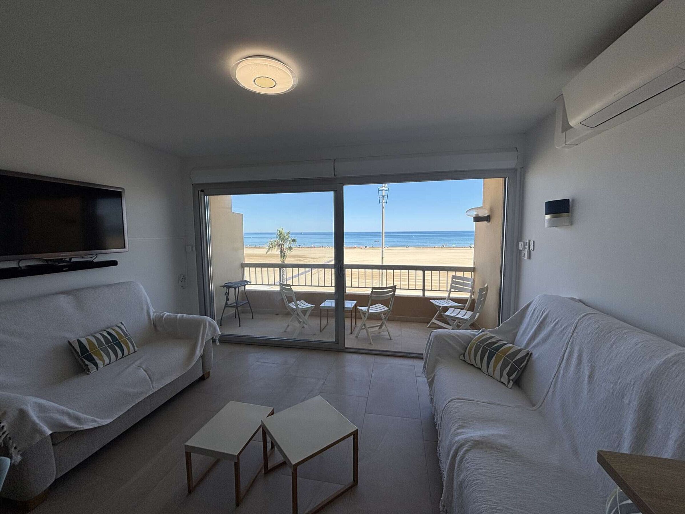 A living room with a TV, two sofas, a small table, and a balcony with a view of the beach.