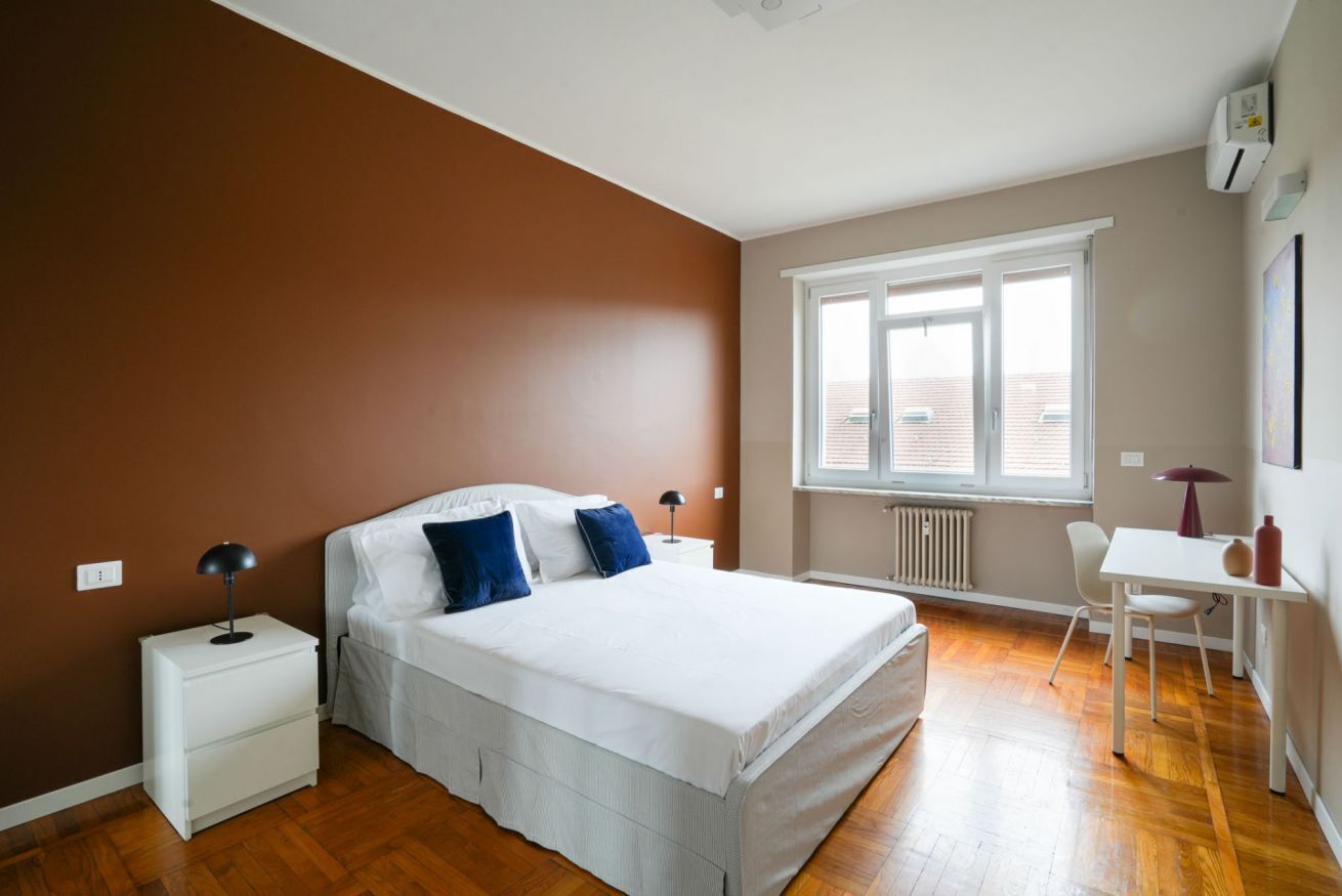 A bedroom with a bed, bedside tables, a window, a radiator, and a desk with a chair.
