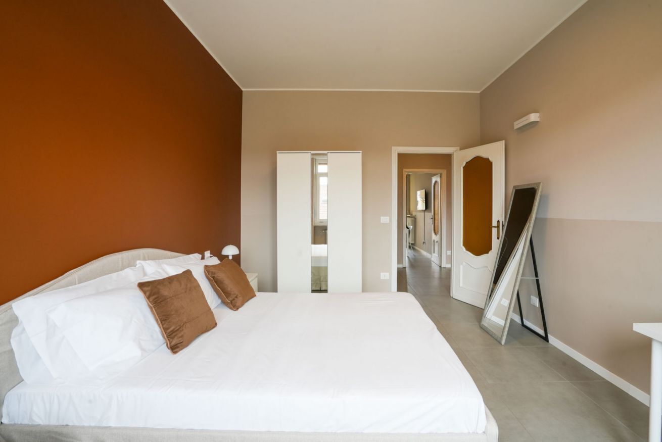 A bedroom with a bed, pillows, a nightstand, a mirror, and a door leading to another room.