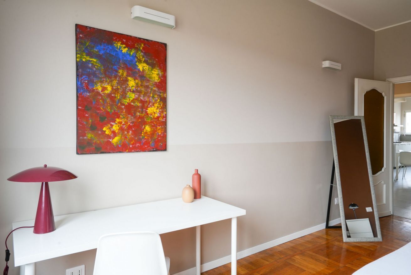 A room with a white desk, a red lamp, a colorful abstract painting, and a large mirror.