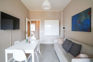 A modern living room with a white sofa, a dining table with chairs, a flat-screen TV mounted on the wall, and a large abstract painting. - Torino Centro | Casa Regina (Torino)