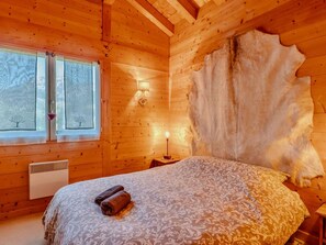 A wooden cabin room with a bed, a window with curtains, a wall-mounted lamp, and a fur rug. - Cozy chalet near slopes - 8 guests (Le Grand-Bornand)