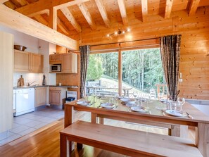 Interior de una cabaña de madera con cocina, mesa de comedor y vistas a un bosque.