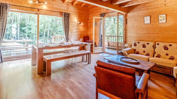 Interior de una cabaña de madera con comedor, sofá y ventanas amplias.