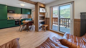 Une cuisine moderne avec des meubles de couleur verte, un coin repas avec une table et des chaises, et une grande porte-fenêtre coulissante donnant sur un balcon avec vue sur un paysage montagneux.